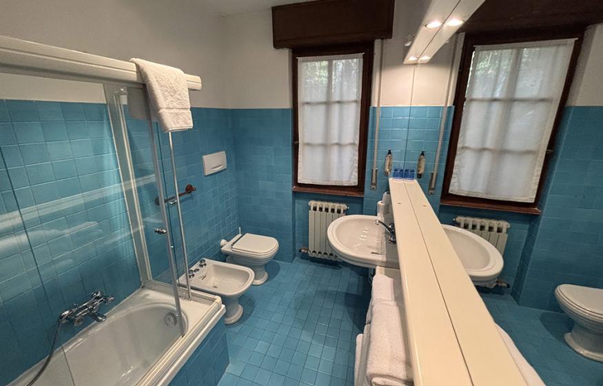 Bagno con piastrelle blu, vasca, bidet e due finestre con tendine.
