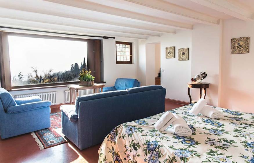 Camera luminosa con letto floreale, divani blu e ampia finestra con vista panoramica.